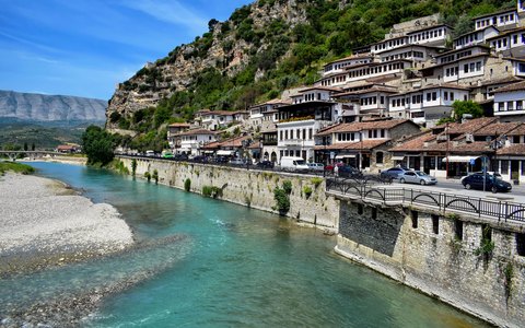 Rows of white Ottoman houses with many windows climbing a green hillside above a turquoise river, under a blue sky