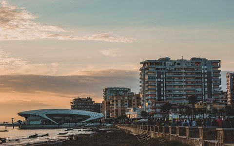 Durrës seafront at sunset, with apartment blocks and palm trees along the promenade and a curved modern building on the shore