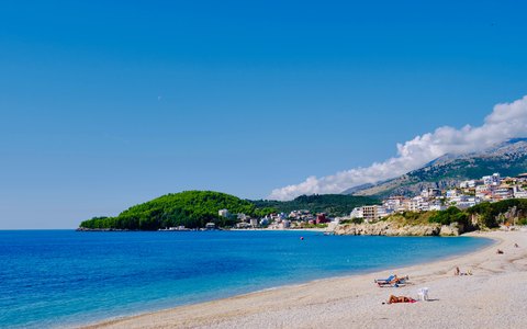 A pebble beach curving along clear blue water towards the white houses of Himarë and a green headland, with mountains and a deep blue sky behind