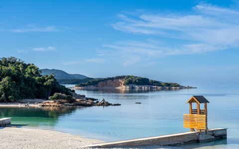 A white sand beach with a wooden lifeguard tower, clear shallow water and green wooded islets under a blue sky