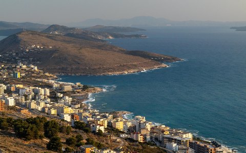 Aerial view of a seaside town along a blue bay, with a brown headland and distant islands in warm evening light