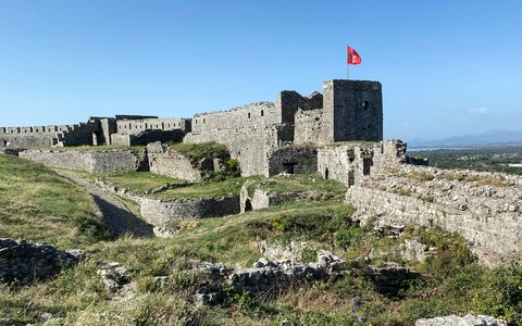 Stone castle walls and a tower flying the Albanian flag on a grassy hilltop under a blue sky