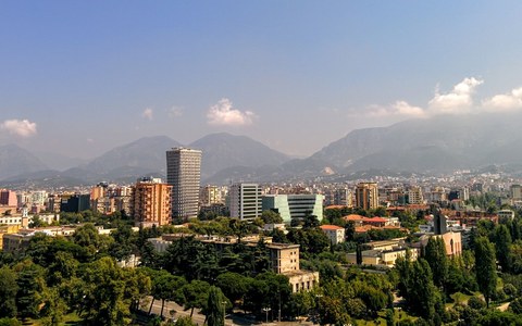 Central Tirana from above, with a tall patterned tower among apartment blocks and parkland, and hazy mountains under a blue sky