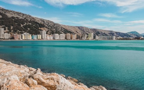 Clear turquoise sea with pale rocks in front and a line of seafront buildings below bare mountains, under a blue sky with thin cloud