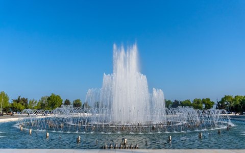 A tall fountain spraying white water in a round pool, with trees around it under a deep blue sky