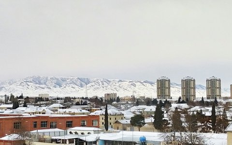 Snow-covered rooftops and low buildings of Ganja, with three tall apartment towers and snowy mountains on the horizon