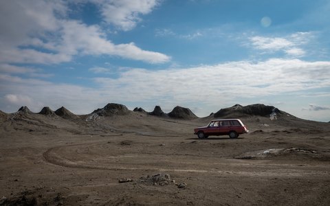Roter Oldtimer, geparkt zwischen den grauen Schlammvulkankegeln von Qobustan unter blauem Himmel mit weißen Wolken