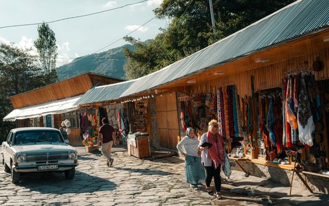 Craft stalls hung with carpets and textiles along a cobbled street in Lahij, with an old white car parked and visitors walking past