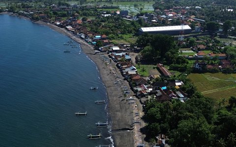Aerial view along the dark sand of Lovina Beach, with outrigger boats in the shallows and village roofs and fields behind