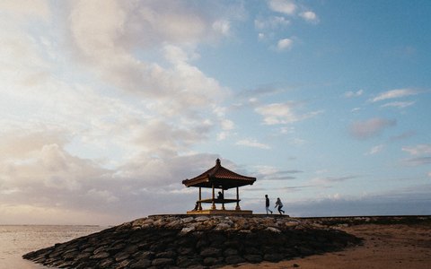 A traditional Balinese gazebo on a stone point beside the calm sea at Sanur, under a soft blue sky