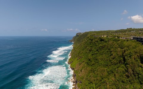 Luftaufnahme der oben grün bewachsenen Klippen von Uluwatu, die steil ins türkisfarbene Meer mit weißer Brandung abfallen