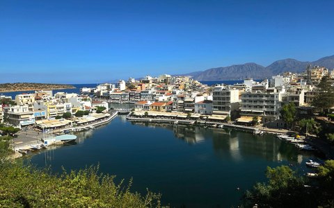 Whitewashed buildings around a calm harbour at Agios Nikolaos, with mountains across the bay under a clear blue sky
