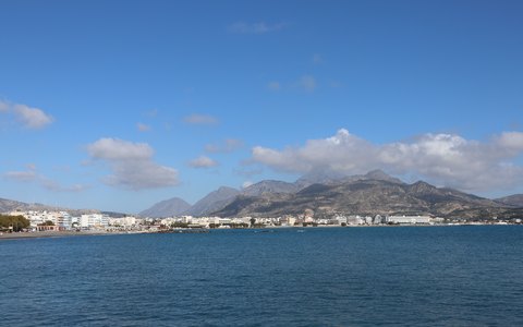 The town of Ierapetra along the shore below mountains, with blue sea in front and a blue sky with white clouds