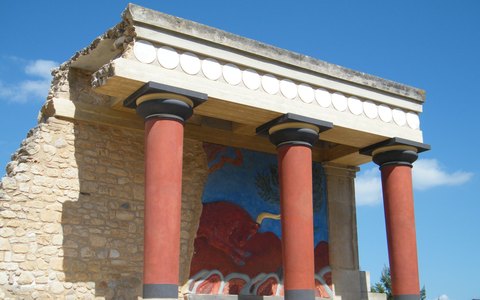 Red Minoan columns and a bull fresco on a restored part of the Knossos palace, under a deep blue sky