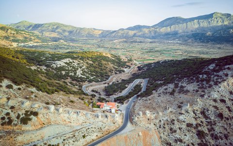 Berge rund um die grünen und braunen Ackerflächen der Lasithi-Hochebene, mit einer Straße, die sich den Hang hinaufschlängelt