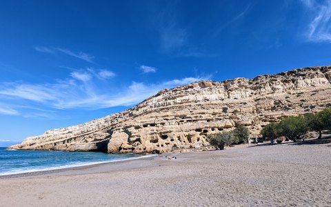A pebble beach below pale cliffs dotted with caves at Matala, under a blue sky with white clouds