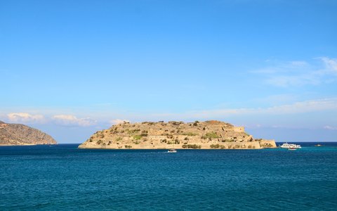 The fortified island of Spinalonga in a deep blue sea, with a hill behind under a clear sky