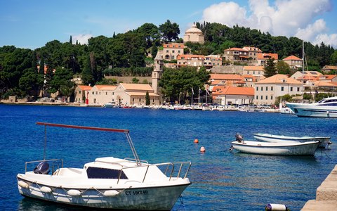 Small boats moored in a blue harbour below Cavtat's stone houses and green hillside, under a blue sky with a few clouds
