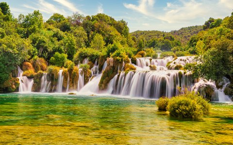 Wide white cascades pouring into a green pool, framed by lush trees and a blue sky with white clouds