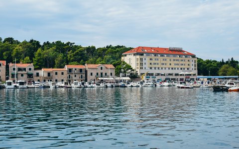 Boats in a calm harbour in front of stone waterfront buildings and a large hotel, with green trees and a blue sky