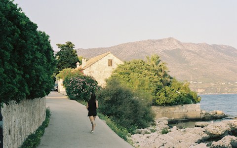 A seafront path past a stone house and green gardens at Orebić, with a bare mountain rising above the sea behind