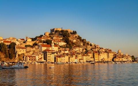 Old stone houses stacked up a hill above the water at Sibenik, glowing in golden evening light under a blue sky