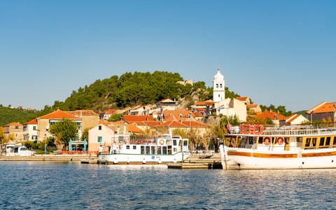 Boats moored along the Skradin waterfront below a hill with a church tower and red-roofed houses, under a clear blue sky