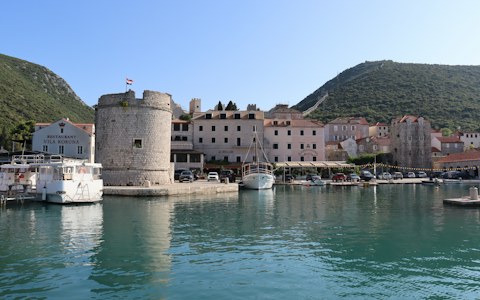 A round stone tower, boats and pastel houses on the harbour at Mali Ston, with green hills and the old town walls behind