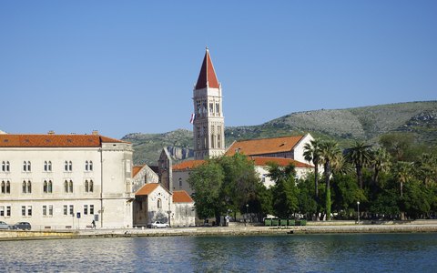 Stone buildings and the cathedral bell tower on the Trogir waterfront, with green hills behind under a blue sky