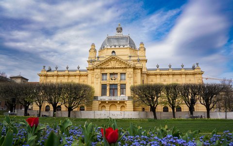 A yellow Art Nouveau pavilion with a glass dome behind spring flower beds and trees, under a blue sky with white clouds