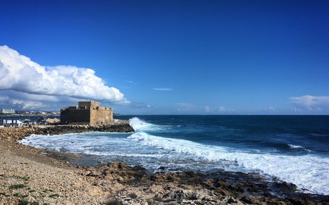 The squat stone Paphos Castle at the end of the harbour, with waves breaking on the rocks and white clouds in a blue sky