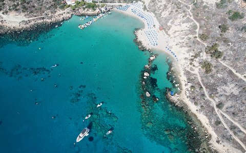 Aerial view of a curved sandy bay with turquoise water and boats, edged by pale rocky ground