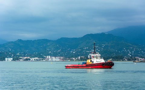A red tugboat crossing Batumi harbour, with the city along the shore and green mountains behind