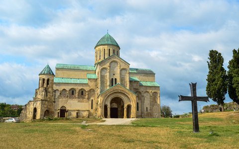 The stone Bagrati Cathedral on a green hilltop with a tall cypress, under a blue sky with white clouds