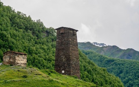 A tall medieval stone tower on a green hillside above Mestia, with forested mountains behind