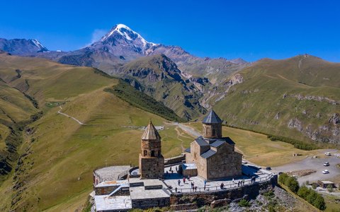 The stone Gergeti Trinity Church on a green ridge with snow-capped Mount Kazbek behind under a deep blue sky