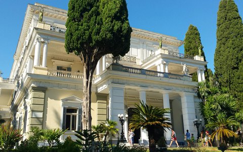 The white neoclassical Achilleion palace behind tall cypress trees and palms, under a blue sky