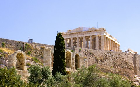 The Parthenon on top of the Acropolis rock above trees and old walls, under a clear blue sky