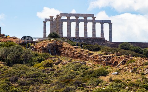 The marble columns of the Temple of Poseidon on a green rocky hilltop, with white clouds in a blue sky