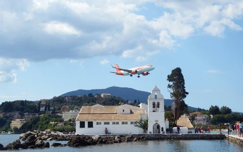 Verkehrsflugzeug im Tiefflug über einem weißen Kloster auf einem Damm am Meer auf Korfu, dahinter grüne Hügel