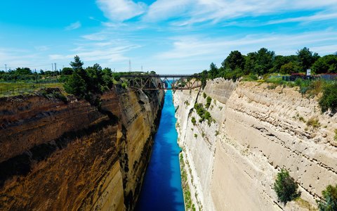 The narrow Corinth Canal cut deep between steep rock walls, with a bridge across it and a blue sky with white clouds