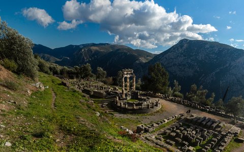 The ancient ruins of Delphi on a green slope below mountains, with white clouds in a blue sky