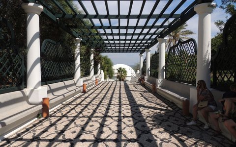 A white-columned pergola walkway with a black and white pebble-mosaic floor, a white dome at the far end and people sitting in the shade
