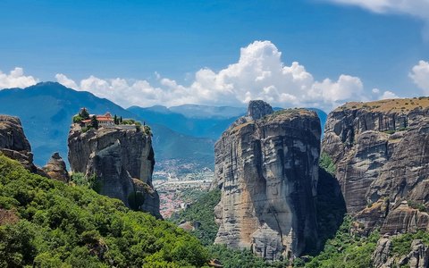 Tall grey rock pillars with monasteries on their tops, green valleys and mountains behind, under a blue sky with white clouds