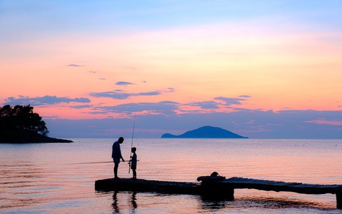 A father and son fishing from a small pier at Neos Marmaras, silhouetted against a pink sunset over the sea and islands