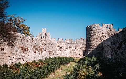 Massive stone walls and a round tower of the medieval town of Rhodes, with plants in front and a blue sky