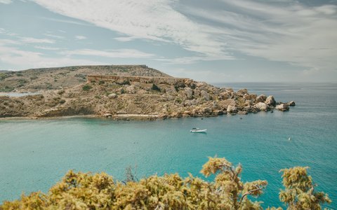 Turquoise water below pale limestone cliffs at Golden Bay, with green scrub in front and a cloudy sky