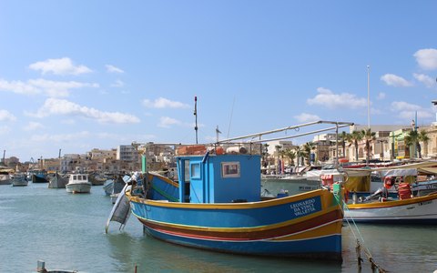 Painted wooden fishing boats moored in Marsaxlokk harbour, with the village waterfront behind under a blue sky with white clouds