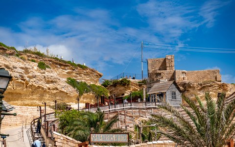 The wooden houses of Popeye Village on a rocky shore above the sea, with palm trees and a blue sky with white clouds