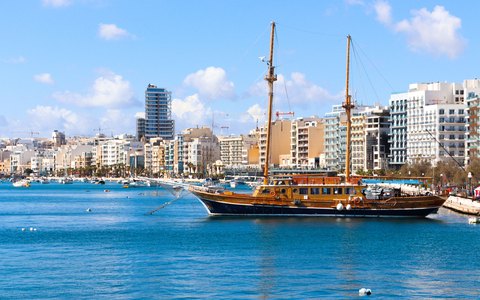 A wooden sailing ship moored in calm blue water in front of the Sliema waterfront, under a blue sky